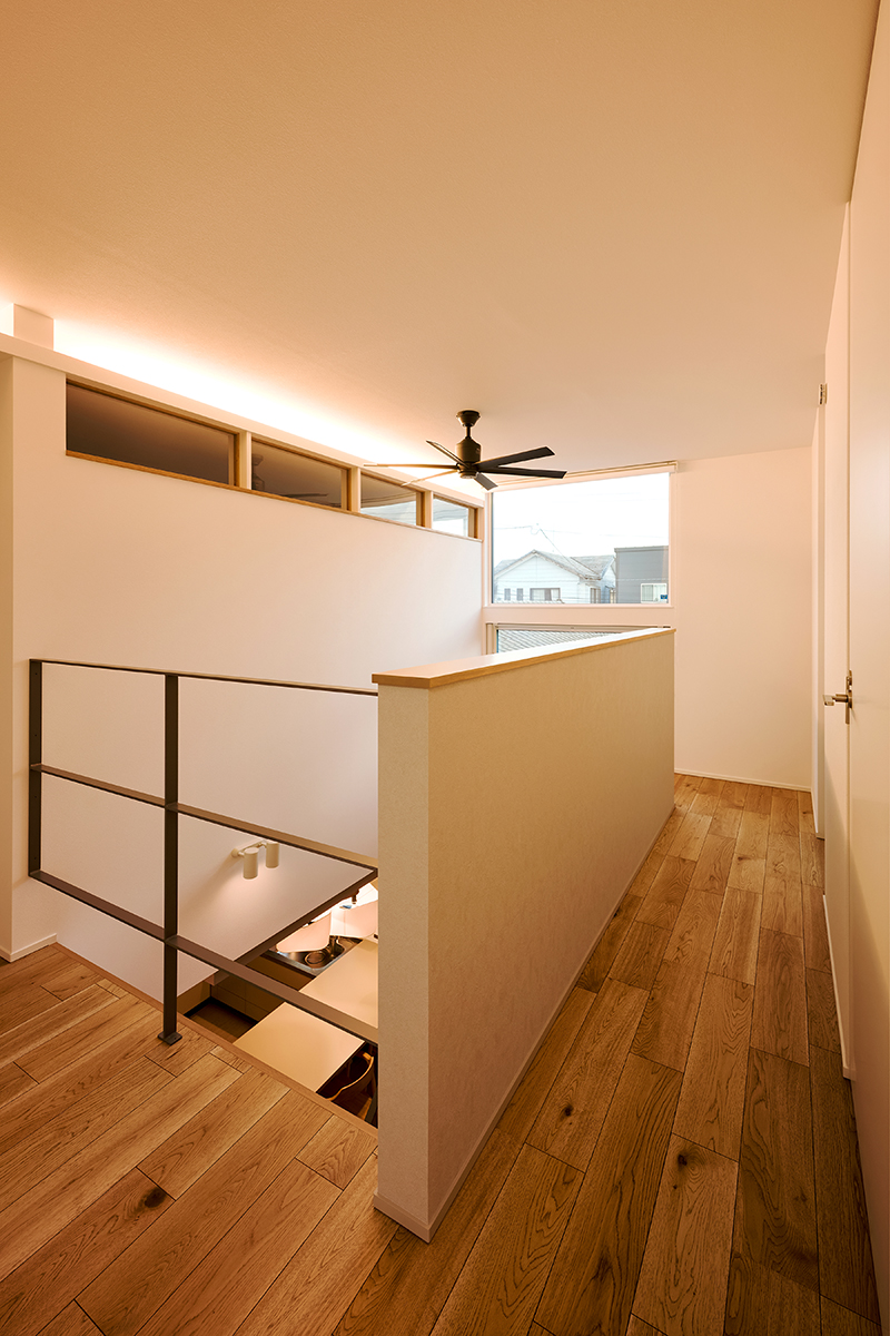2階ホールからも吹抜けの開放感を味わえる。大きなFIX窓からの景色や間接照明、寝室の室内窓が印象的。黒のシーリングファンやアイアン手すりが空間を引き締める